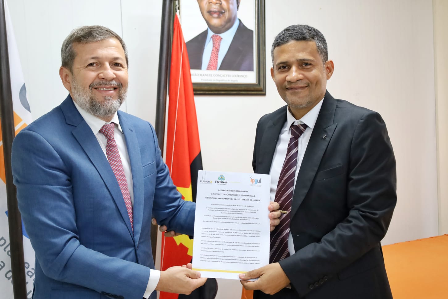 A fotografia está em plano médio e mostra o vice-prefeito de Fortaleza, Élcio Batista, à esquerda; e o diretor do Instituto de Planejamento e Gestão Urbana de Luana, Oswaldo Fortes, à direita. Os dois posam para a imagem e seguram o documento que simboliza a parceria. Élcio está de terno azul marinho, com gravata vermelha e camisa branca. Oswaldo usa terno preto, com grava camisa branca e gravata listrada. Ao fundo, está uma bandeira de Luanda.  